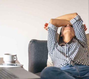 Wanita bersandar di sofa sambil menutupi wajah dengan tangan di depan laptop sebagai tanda tubuh butuh istirahat setelah bekerja seharian.