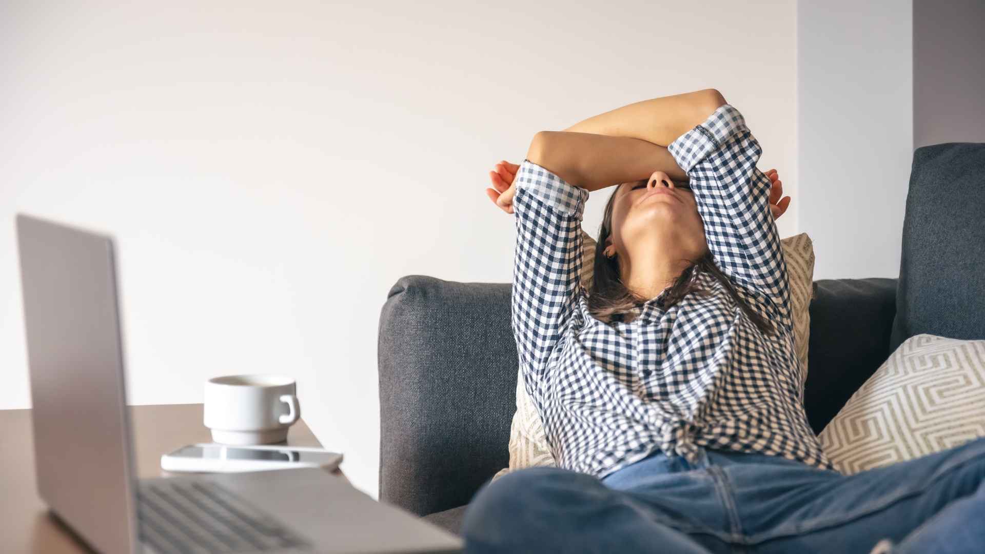 Wanita bersandar di sofa sambil menutupi wajah dengan tangan di depan laptop sebagai tanda tubuh butuh istirahat setelah bekerja seharian.